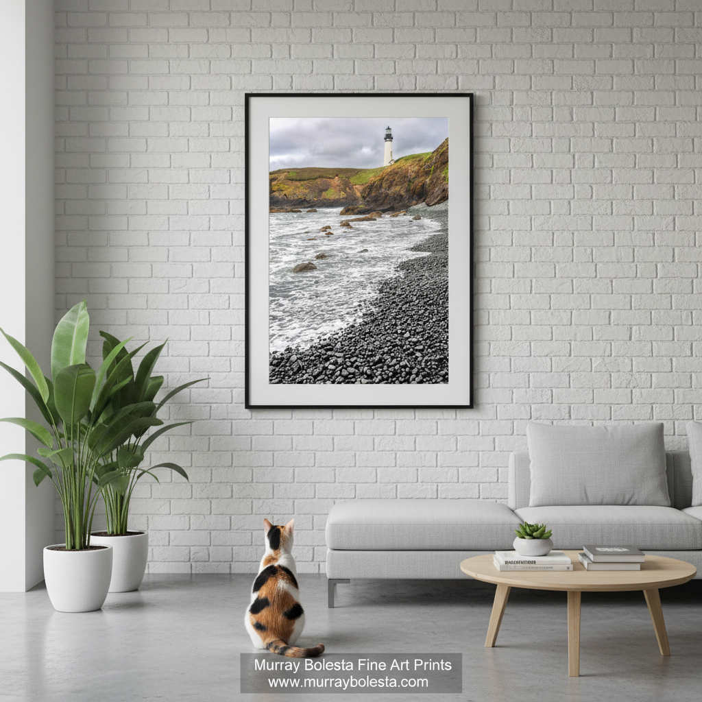 Yaquina Head by Murray Bolesta! A framed vertical art print of a coastal lighthouse scene hangs on a white brick wall above a light gray sofa in a modern living room. A calico cat sits on the floor below the frame, and a small round wooden coffee table is in front of the sofa.