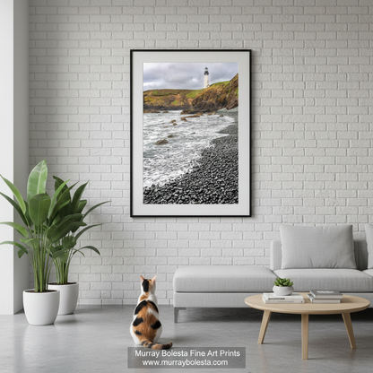 Yaquina Head by Murray Bolesta! A framed vertical art print of a coastal lighthouse scene hangs on a white brick wall above a light gray sofa in a modern living room. A calico cat sits on the floor below the frame, and a small round wooden coffee table is in front of the sofa.